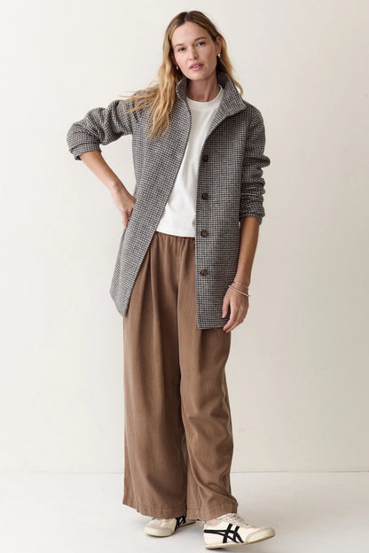 A woman modeling a stylish outfit consisting of a gray and black houndstooth button-up jacket, brown wide-leg trousers, a white t-shirt, and white sneakers with black stripes, set against a plain off-white studio background.