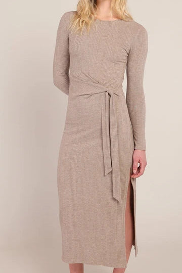 A woman wearing a ribbed knit long-sleeve midi dress in a warm taupe-beige color, featuring a self-tie waist and a side slit.