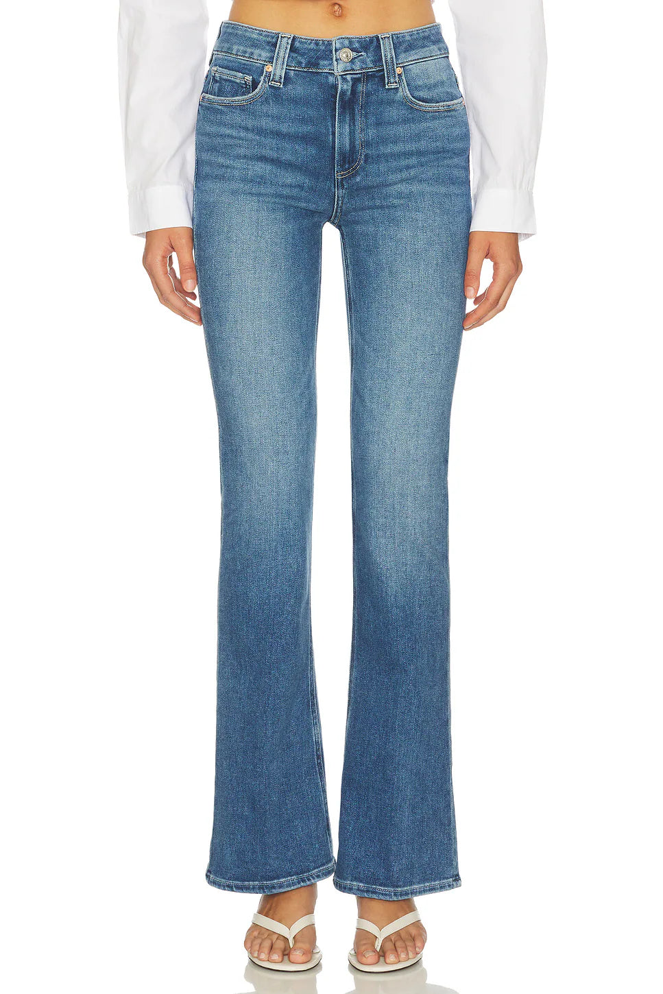 A model wearing a pair of medium-wash blue denim flare jeans, styled with a white long-sleeve shirt and white sandals, shown from the waist down on a white background.