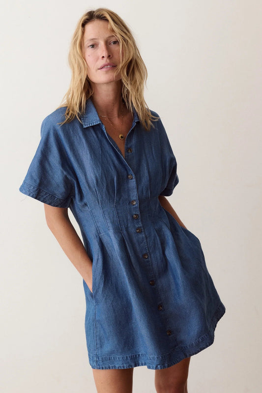 A woman wearing a short-sleeve blue denim shirt dress with a button-down front and side pockets, standing against a plain off-white background.