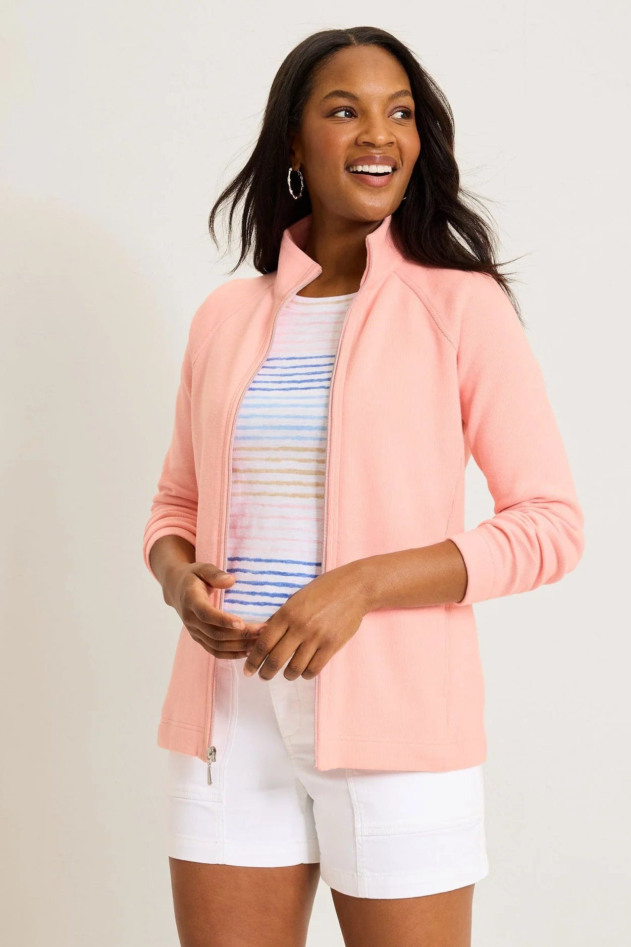 Woman wearing a pink jacket over a striped shirt and white shorts on a plain background