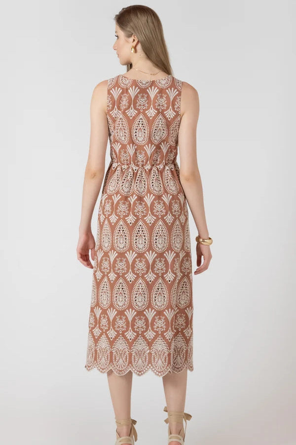 A woman viewed from the back wearing a sleeveless midi dress with an intricate paisley and floral lace pattern in terracotta and beige, paired with beige platform sandals, against a light gray studio background.