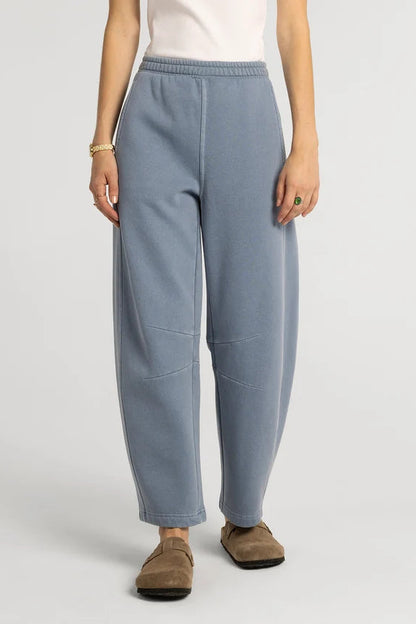 A person wearing loose, wide-leg light blue sweatpants with an elastic waistband, paired with a white top and brown slip-on shoes, photographed against a plain light gray studio background.