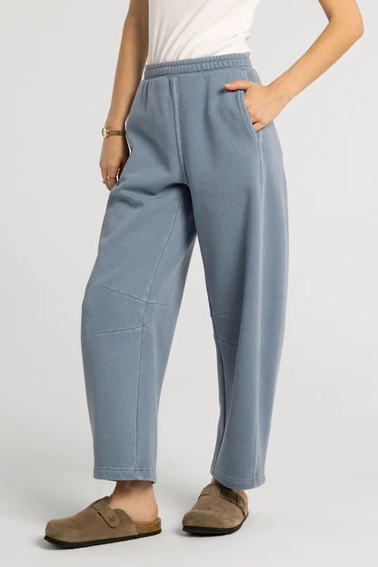 A person wearing light blue wide-leg cropped sweatpants with side pockets, paired with a white top and brown slip-on clogs, photographed against a plain light gray studio background.