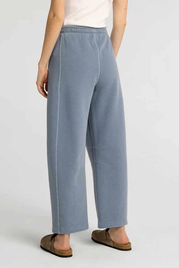 A person wearing wide-leg light blue sweatpants with an elastic waistband, paired with a white top and brown slide sandals, shown from the back against a light gray studio background.