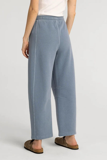 A person wearing wide-leg light blue sweatpants with an elastic waistband, paired with a white top and brown slide sandals, shown from the back against a light gray studio background.