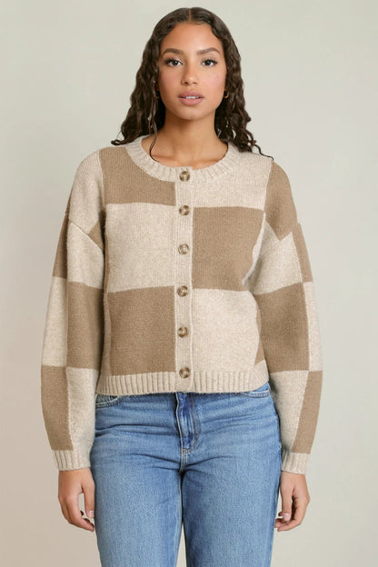 A woman wearing a beige and cream checkered button-up cardigan sweater with blue jeans, standing against a neutral light gray background.