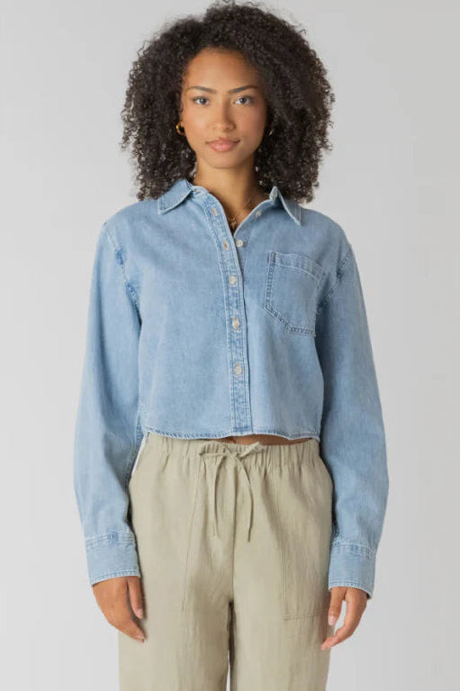 Woman wearing a light blue denim shirt and beige pants against a plain background