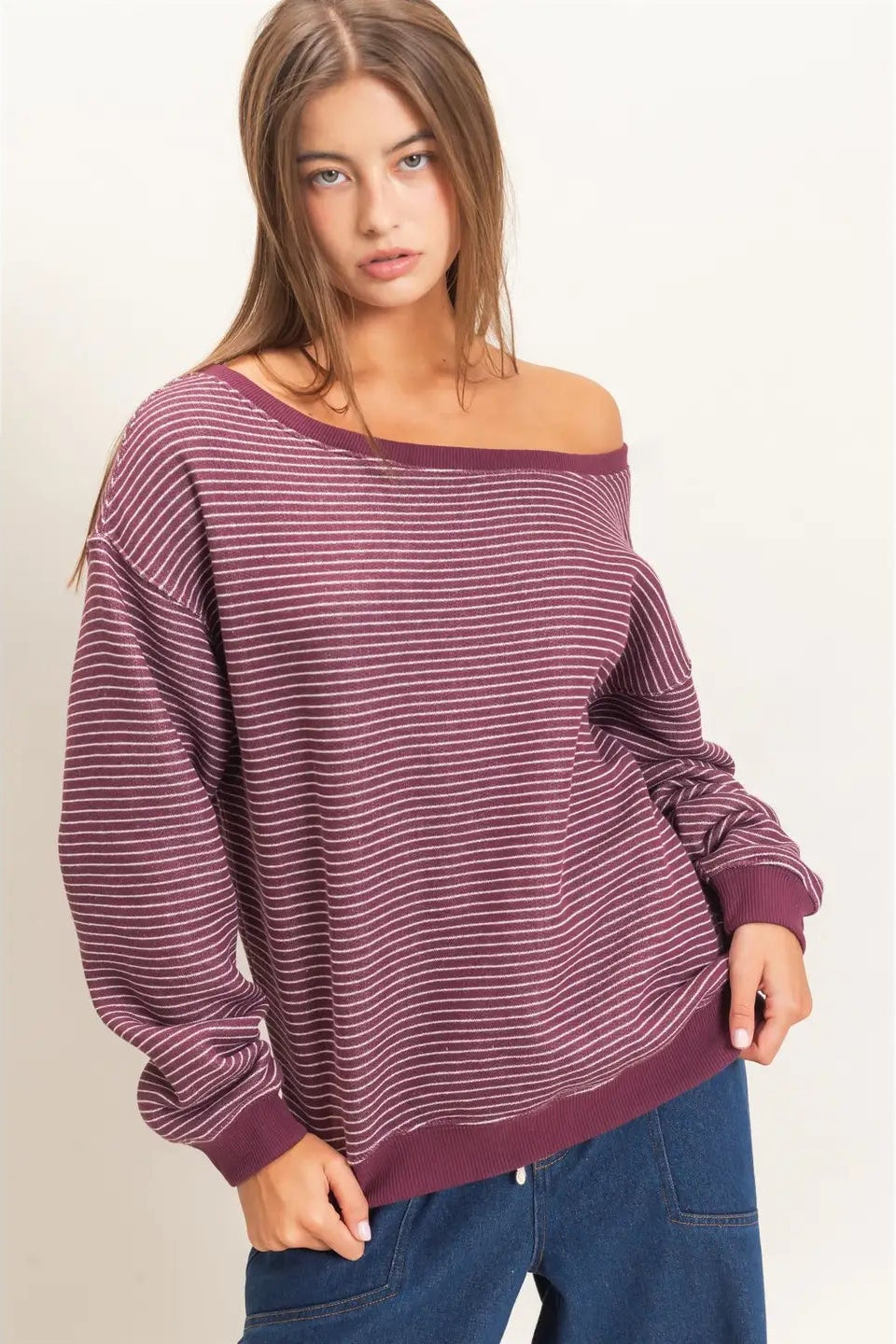 A woman wearing a long-sleeve off-the-shoulder sweater with thin horizontal burgundy and gray stripes, paired with blue jeans, against a plain light background.