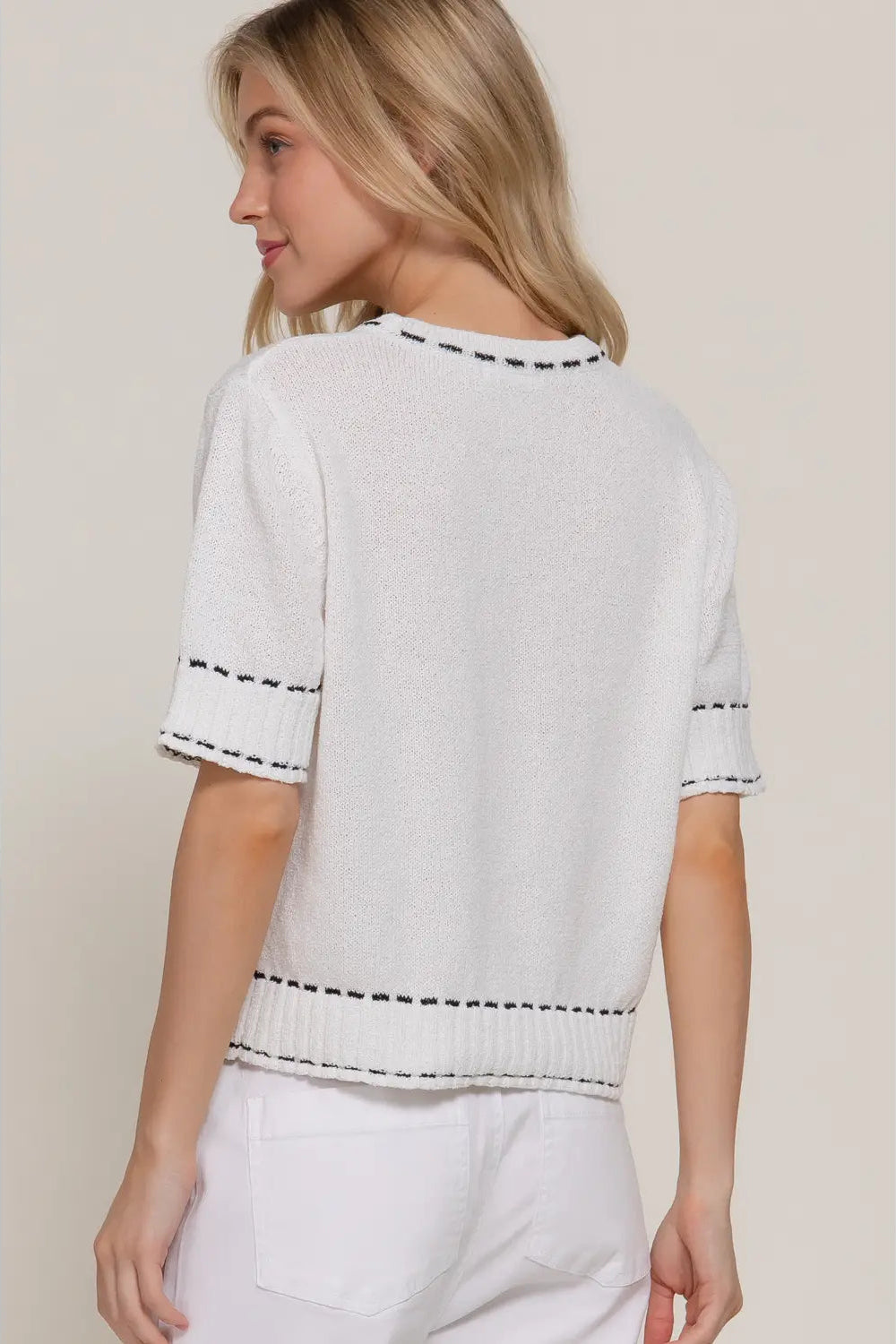 A woman shown from the back wearing a white short-sleeve knit top with black dashed trim details along the neckline, sleeve cuffs, and hem, paired with white pants against a light beige background.