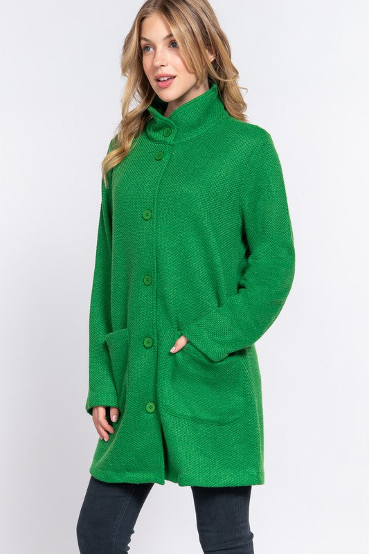 A woman wearing a bright green textured knit coat with a high mock neck, button front, and front patch pockets, paired with dark jeans against a light gray studio background.