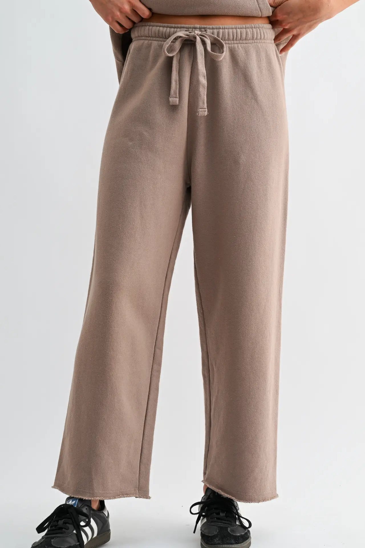 A person wearing loose, wide-leg sweatpants in a muted taupe-brown color, paired with black sneakers, shown from the waist down against a plain light gray studio background.