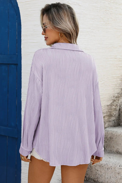 Woman wearing a light purple button-up shirt against a textured white wall.