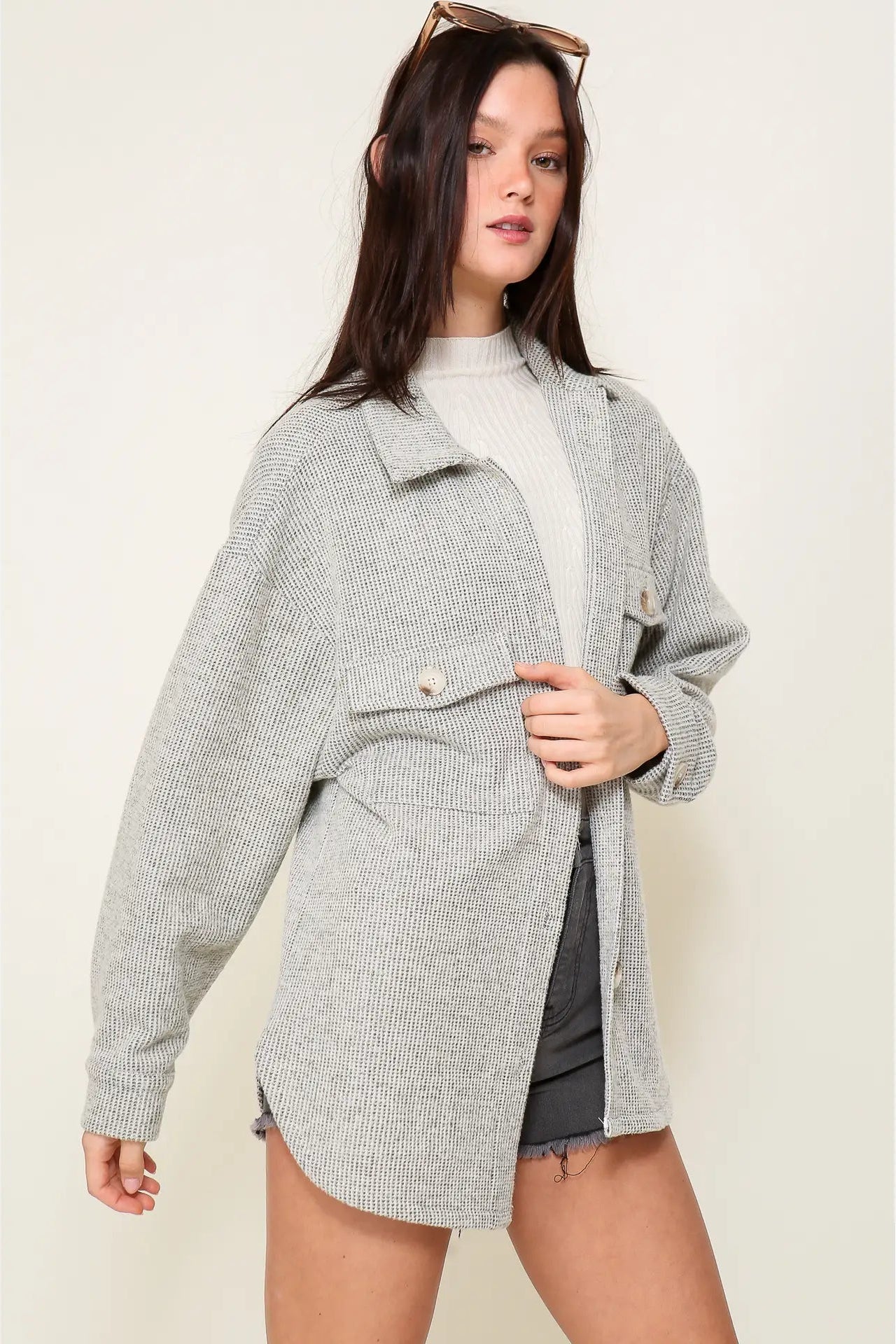 A woman modeling an oversized light gray textured jacket over a cream ribbed turtleneck and dark gray shorts, against a plain beige studio background.