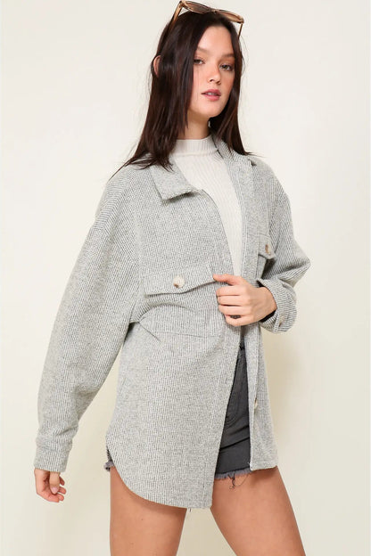 A woman modeling an oversized light gray textured jacket over a cream ribbed turtleneck and dark gray shorts, against a plain beige studio background.