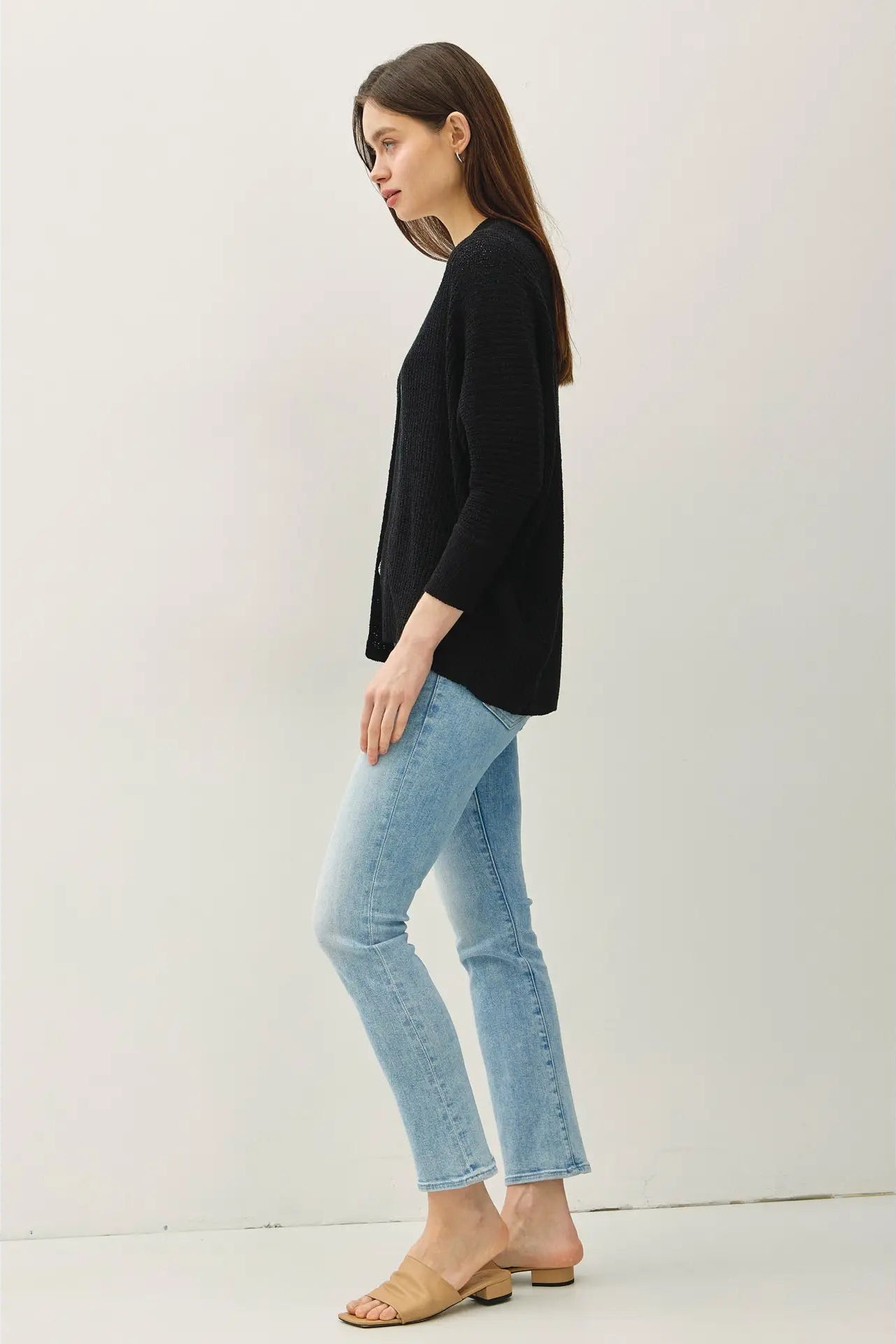 A woman modeling a black three-quarter sleeve knit top with light-wash blue cropped jeans and beige flat sandals, standing in profile against a plain off-white studio background.