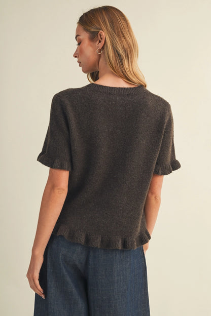 A woman shown from the back wearing a dark brown short-sleeve knit top with a ruffled hem and cuffs, paired with blue jeans, against a plain light beige background.