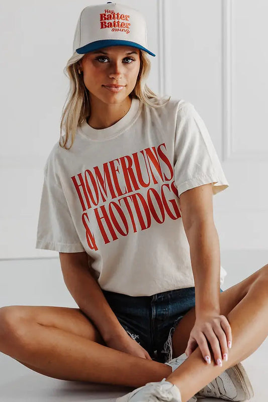 Woman wearing a white t-shirt with 'Homeruns & Hotdogs' text and a cap with red and blue design.
