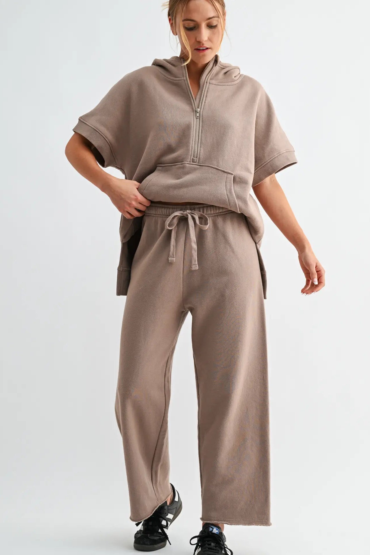 A woman modeling a matching taupe-brown lounge set consisting of a short-sleeve half-zip hoodie and wide-leg sweatpants, paired with black sneakers against a light gray studio background.