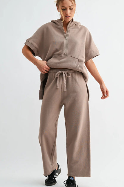 A woman modeling a matching taupe-brown lounge set consisting of a short-sleeve half-zip hoodie and wide-leg sweatpants, paired with black sneakers against a light gray studio background.