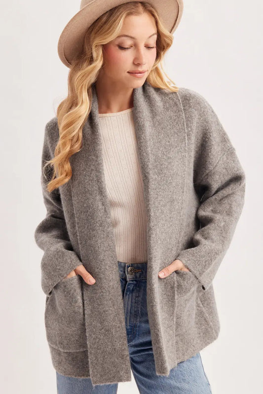 A woman wearing a heather gray open-front cardigan with pockets, a cream ribbed top, blue jeans, and a tan wide-brim hat, photographed against a plain light background.