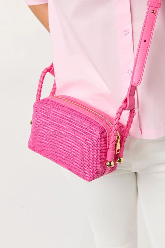 Pink woven handbag with gold accents worn by a person in a pink shirt and white pants on a light background