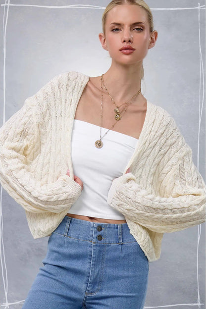 A woman wearing a chunky cream cable-knit cardigan over a white strapless top and blue jeans, posed against a textured gray background with a white hand-drawn border.