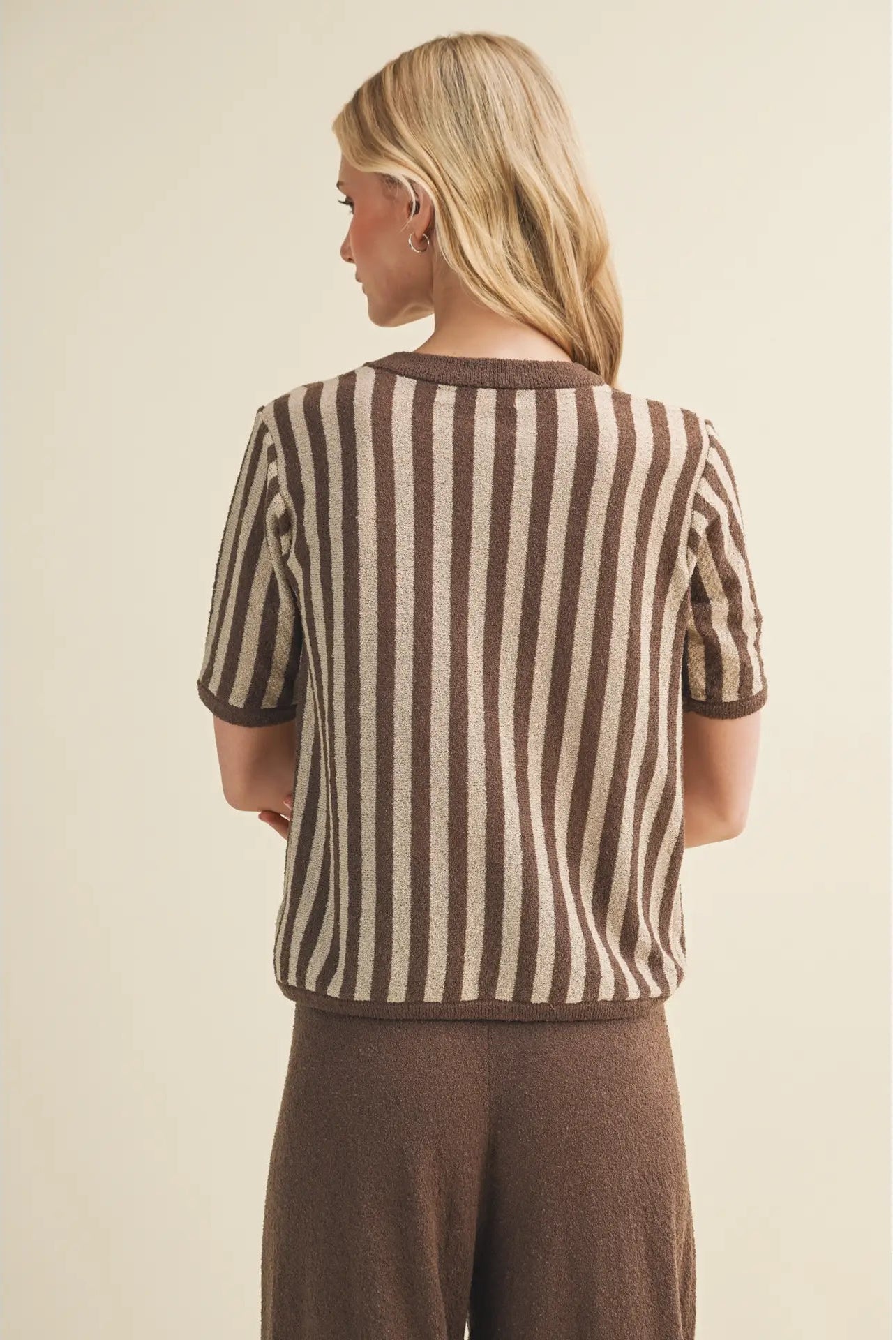A woman shown from the back wearing a short-sleeve knit top with vertical brown and beige stripes, paired with brown pants, against a plain beige studio background.