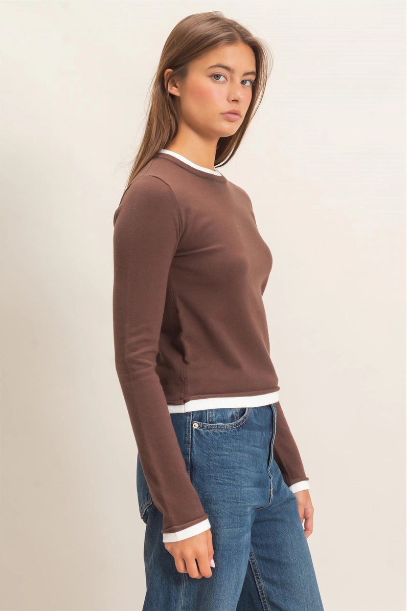 A woman wearing a brown long-sleeve crewneck sweater with white ribbed trim at the collar, cuffs, and hem, paired with blue jeans, shown against a plain light beige studio background.