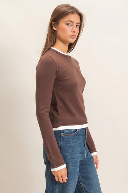A woman wearing a brown long-sleeve crewneck sweater with white ribbed trim at the collar, cuffs, and hem, paired with blue jeans, shown against a plain light beige studio background.