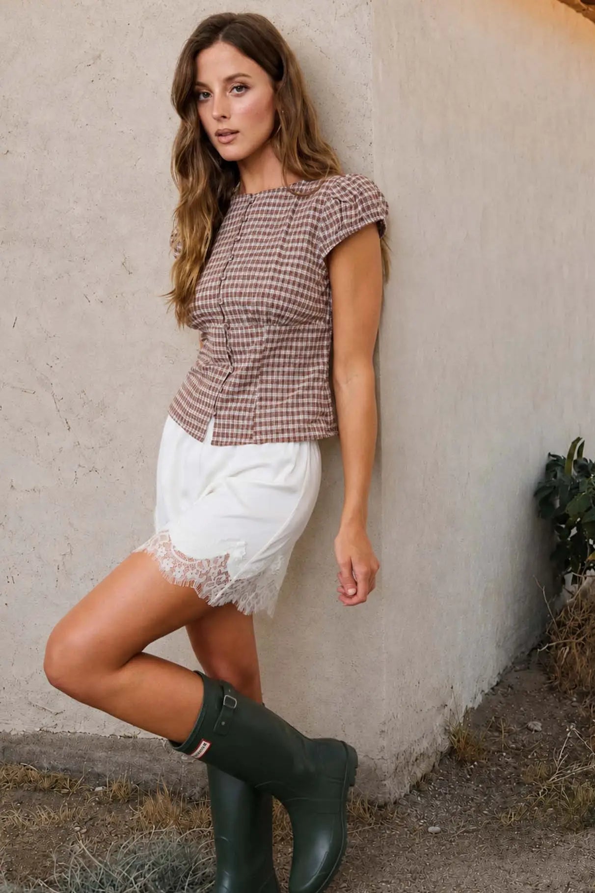 A woman posing outdoors against a stucco wall, wearing a brown and white gingham short-sleeve top, white shorts with a lace hem, and dark green rain boots.