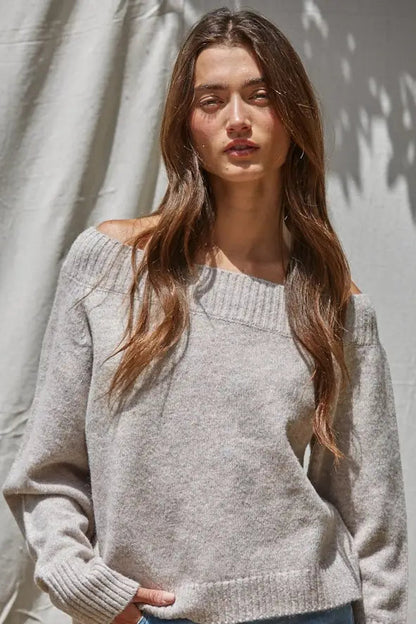 A woman wearing a light heather gray off-the-shoulder knit sweater, standing against a textured light gray wall with soft shadows.
