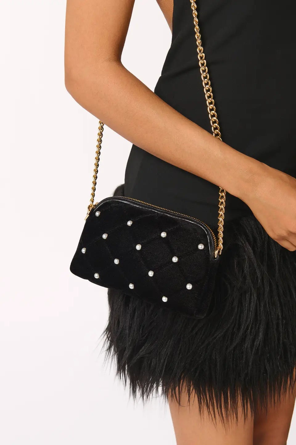 A black quilted velvet crossbody bag with white pearl-like studs and a gold chain strap, shown worn by a model in a black dress against a white background.