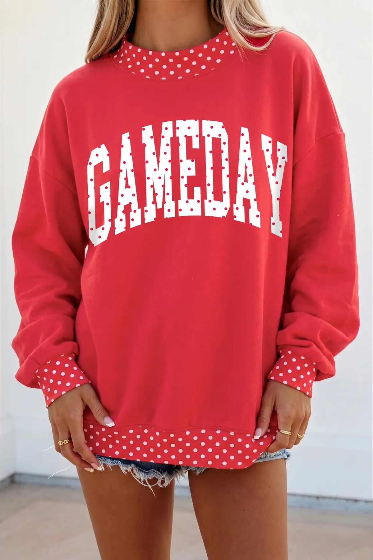 A woman wearing a red crewneck sweatshirt with 'GAMEDAY' printed in white arched letters across the chest. The sweatshirt features white polka-dot trim around the neckline, cuffs, and hem, and is styled with denim shorts.