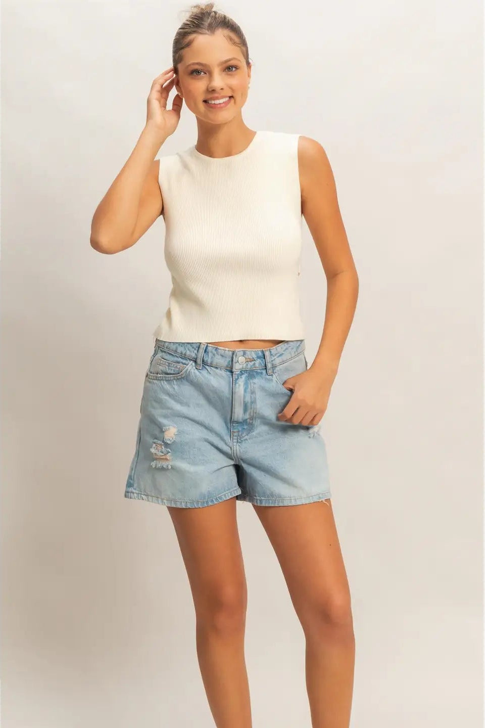 Woman wearing a white sleeveless top and denim shorts on a plain background