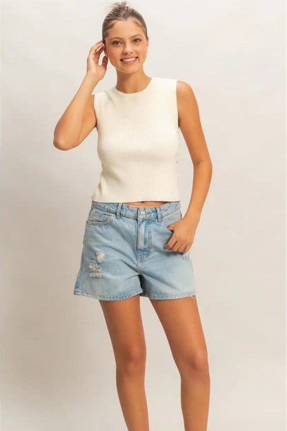 Woman wearing a white sleeveless top and denim shorts on a plain background
