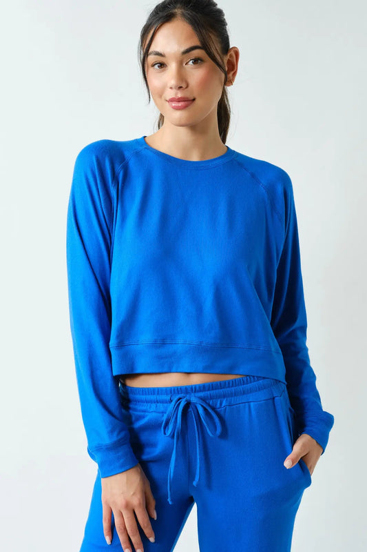 A woman modeling a bright royal blue matching lounge set, featuring a cropped long-sleeve sweatshirt and drawstring sweatpants, against a plain light gray studio background.