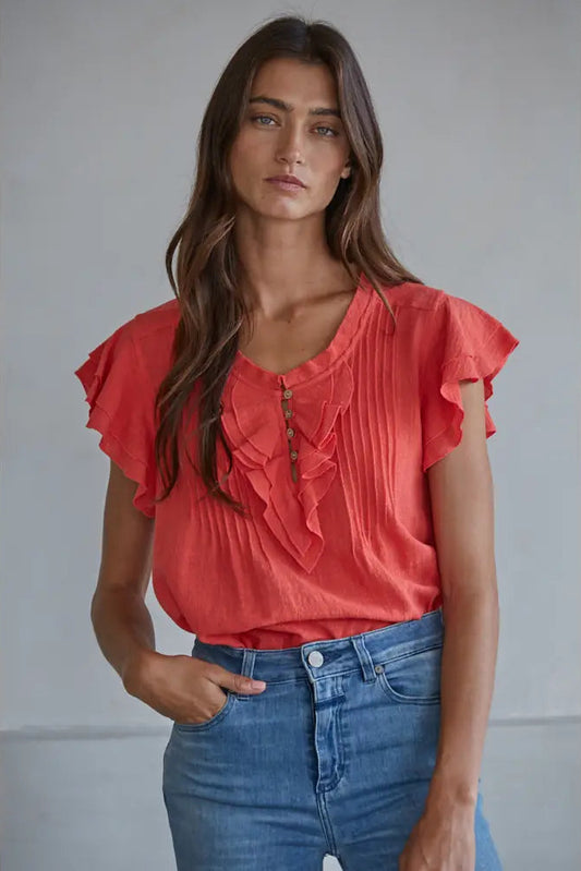 Woman wearing a coral blouse with ruffled sleeves and blue jeans against a gray background