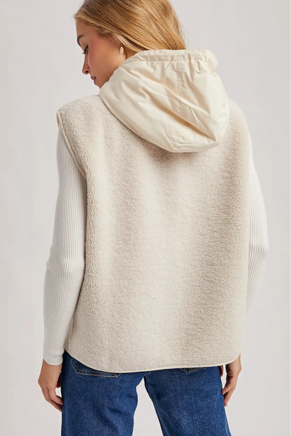 A woman shown from the back wearing a cream-colored, fuzzy hooded sleeveless vest over a white ribbed long-sleeve top and blue jeans, against a plain light gray studio background.