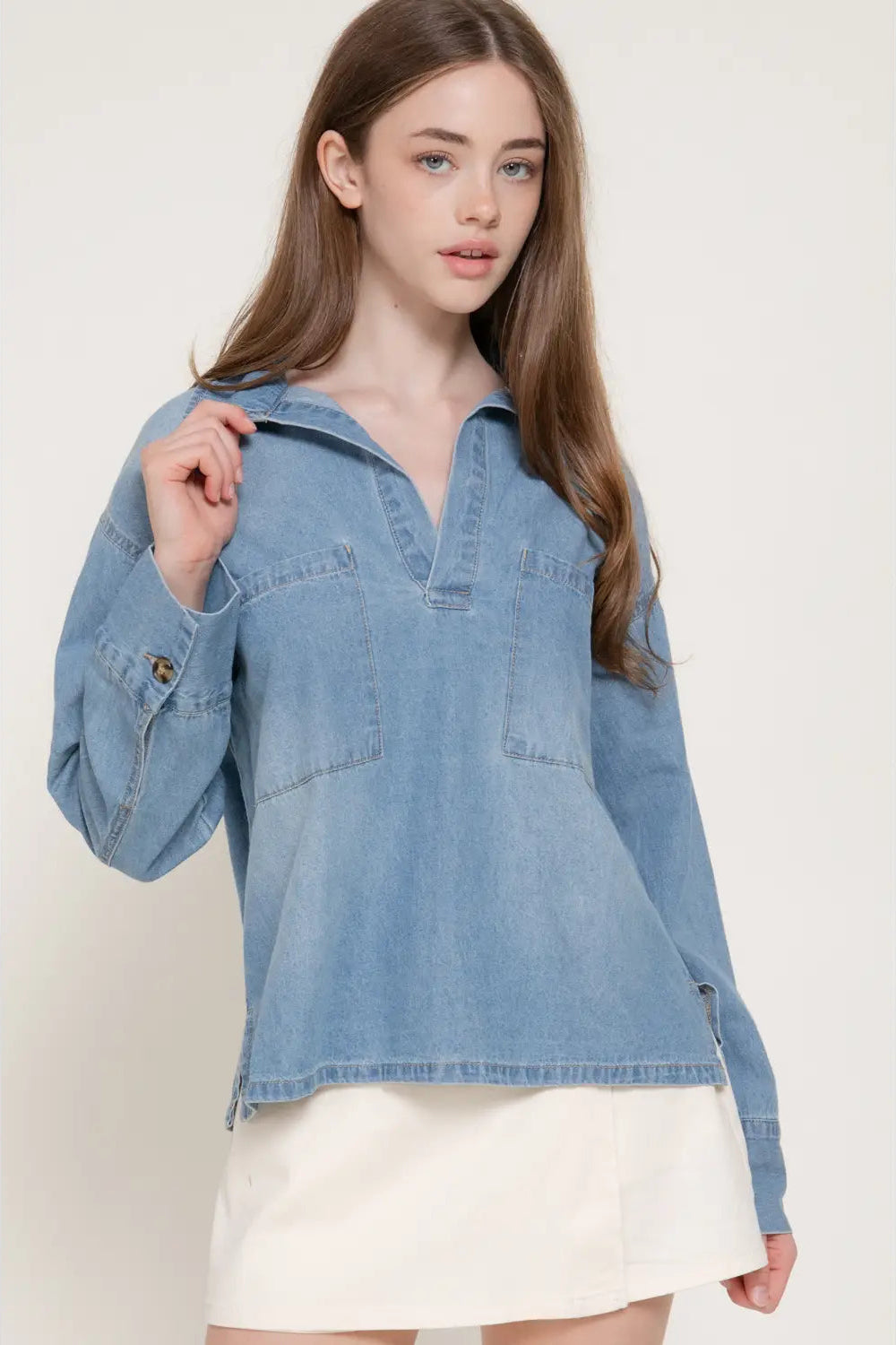 A woman wearing a light blue denim long-sleeve shirt with a split collar and a white skirt, photographed against a plain light beige studio background.