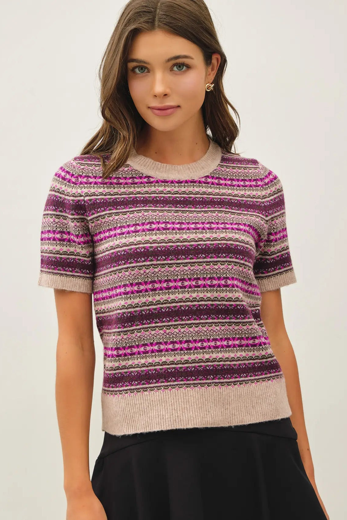 A woman wearing a short-sleeve crewneck knit sweater with horizontal Fair Isle style stripes in purple, pink, and beige, paired with a black skirt against a plain light background.