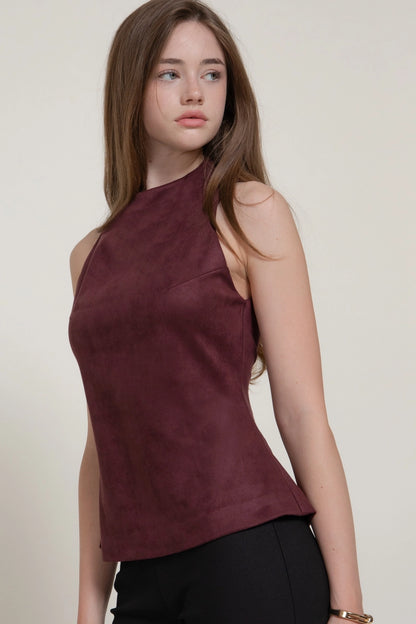 A woman wearing a sleeveless, high-neck, peplum-style top in a deep burgundy or maroon color, paired with black pants, against a plain light beige background.