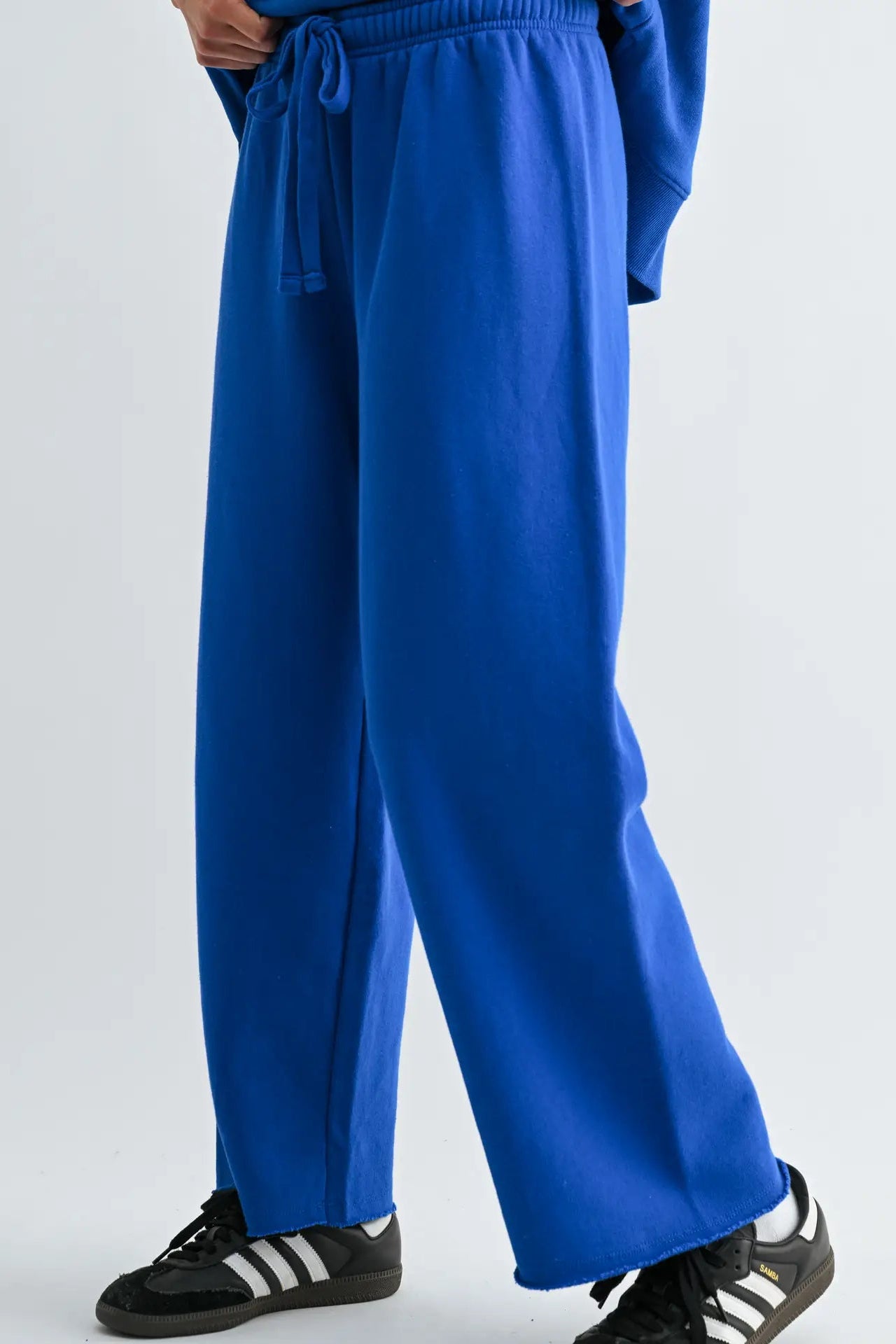A person wearing bright royal blue wide-leg sweatpants with an elastic drawstring waist, paired with black and white sneakers, shown from the waist down against a light gray studio background.