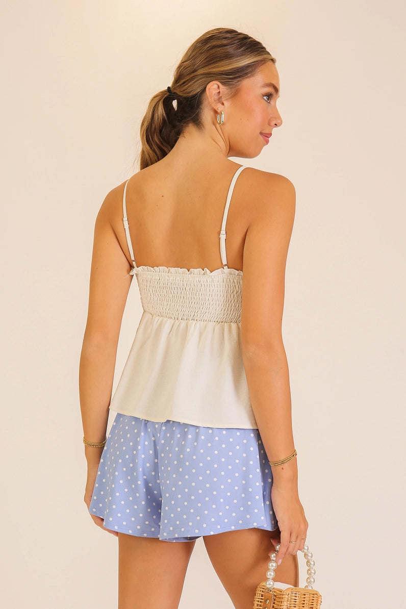 A woman shown from the back wearing a cream-colored smocked camisole top and light blue polka dot shorts, holding a small woven basket against a solid beige background.