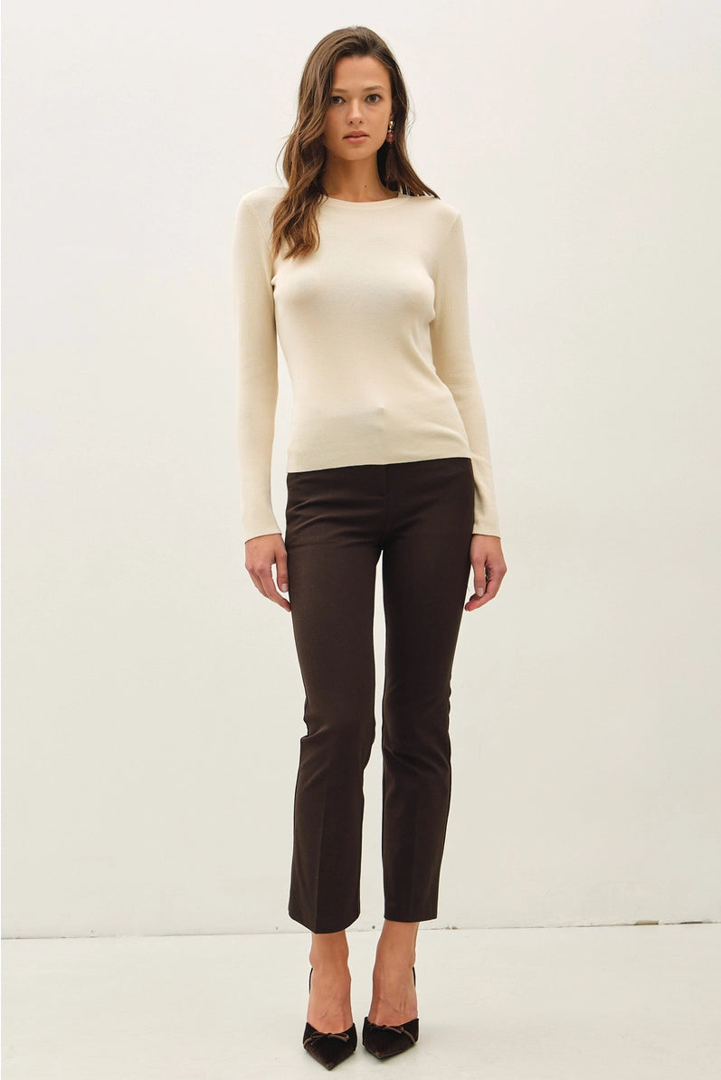 A woman modeling a cream long-sleeve knit sweater and dark brown cropped trousers with black pointed-toe heels, standing against a plain off-white studio background.