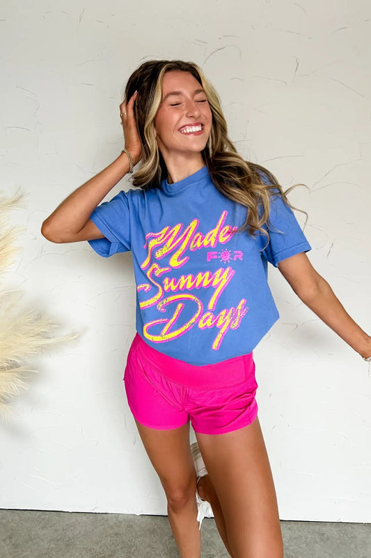 Woman wearing a blue t-shirt with colorful text and pink shorts, standing against a white wall.