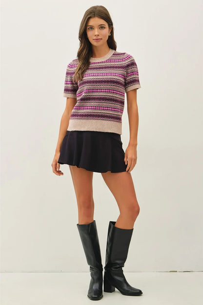 A woman modeling a short-sleeve knit sweater with horizontal stripes in pink, purple, and beige, paired with a black mini skirt and black knee-high boots, against a plain off-white studio background.