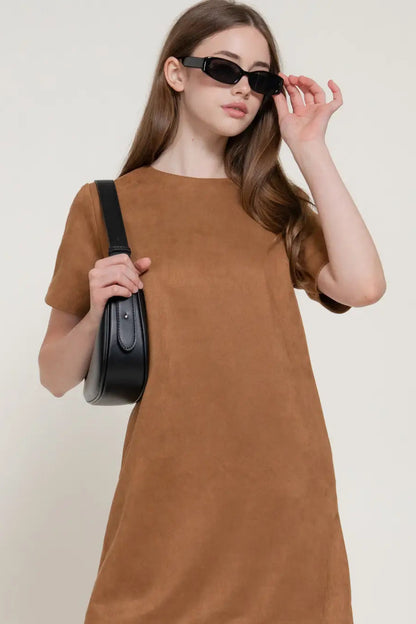 A woman wearing a brown short-sleeve dress and black sunglasses, holding a black shoulder bag against a plain light beige background.