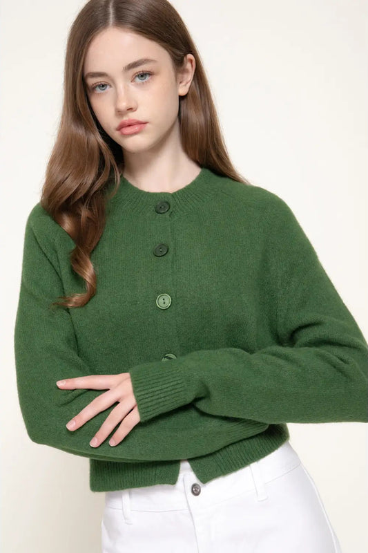 A woman wearing a dark green knit cardigan sweater with dark buttons and white pants, photographed against a plain light beige studio background.