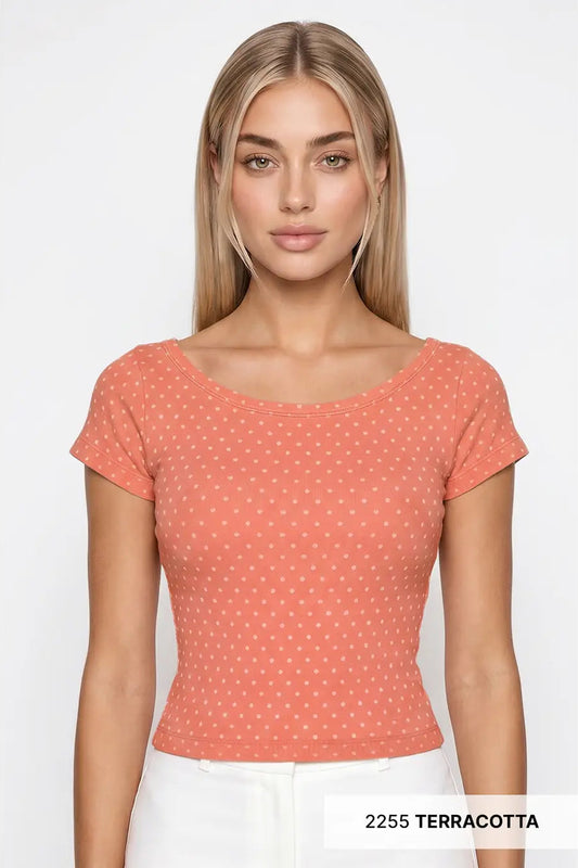 A woman modeling a terracotta short-sleeve scoop-neck top with a small white geometric pattern, paired with white pants against a light gray background.